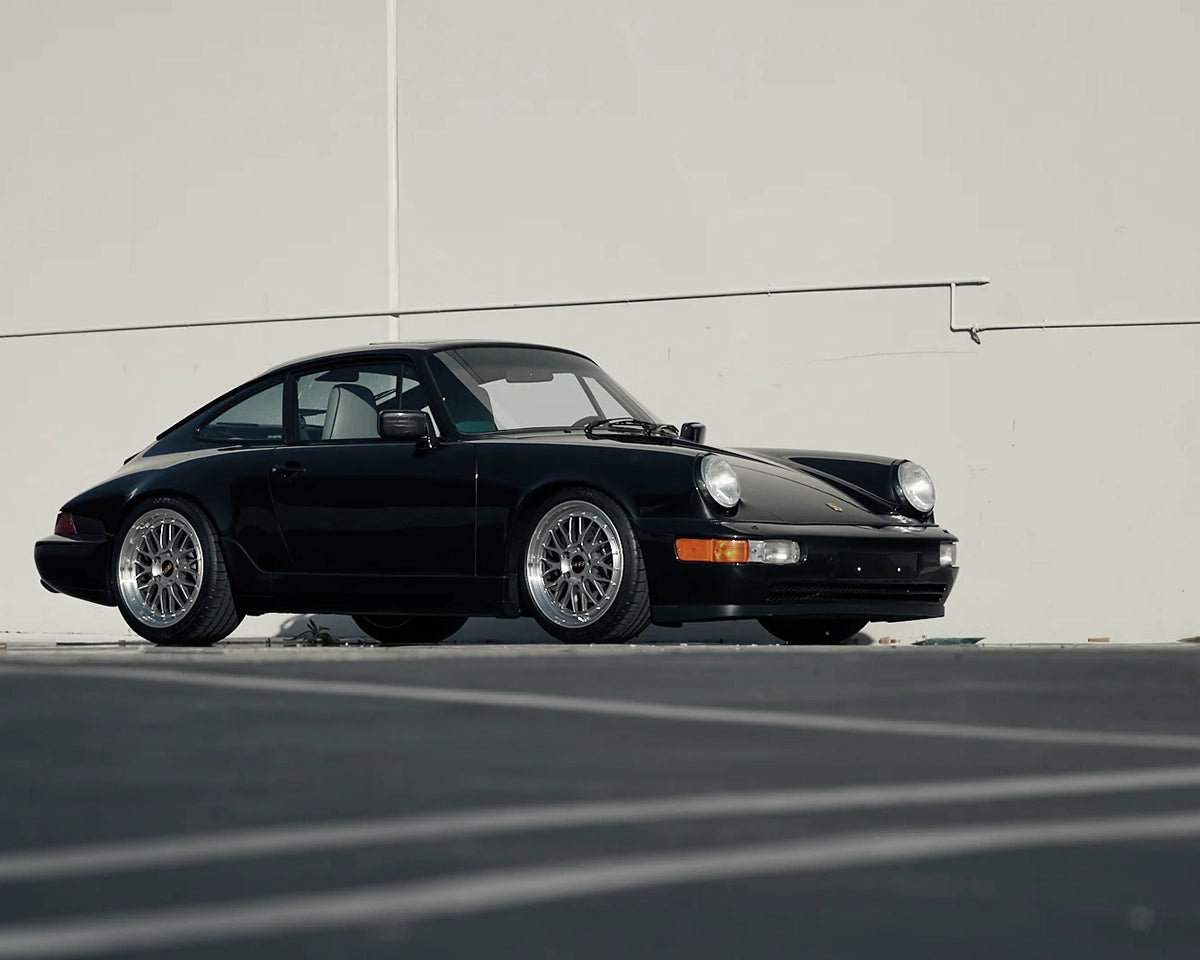 Black narrow-body Porsche 911 Carrera 964 fitted with silver BBS LM Le Mans wheels, front three-quarter view.
